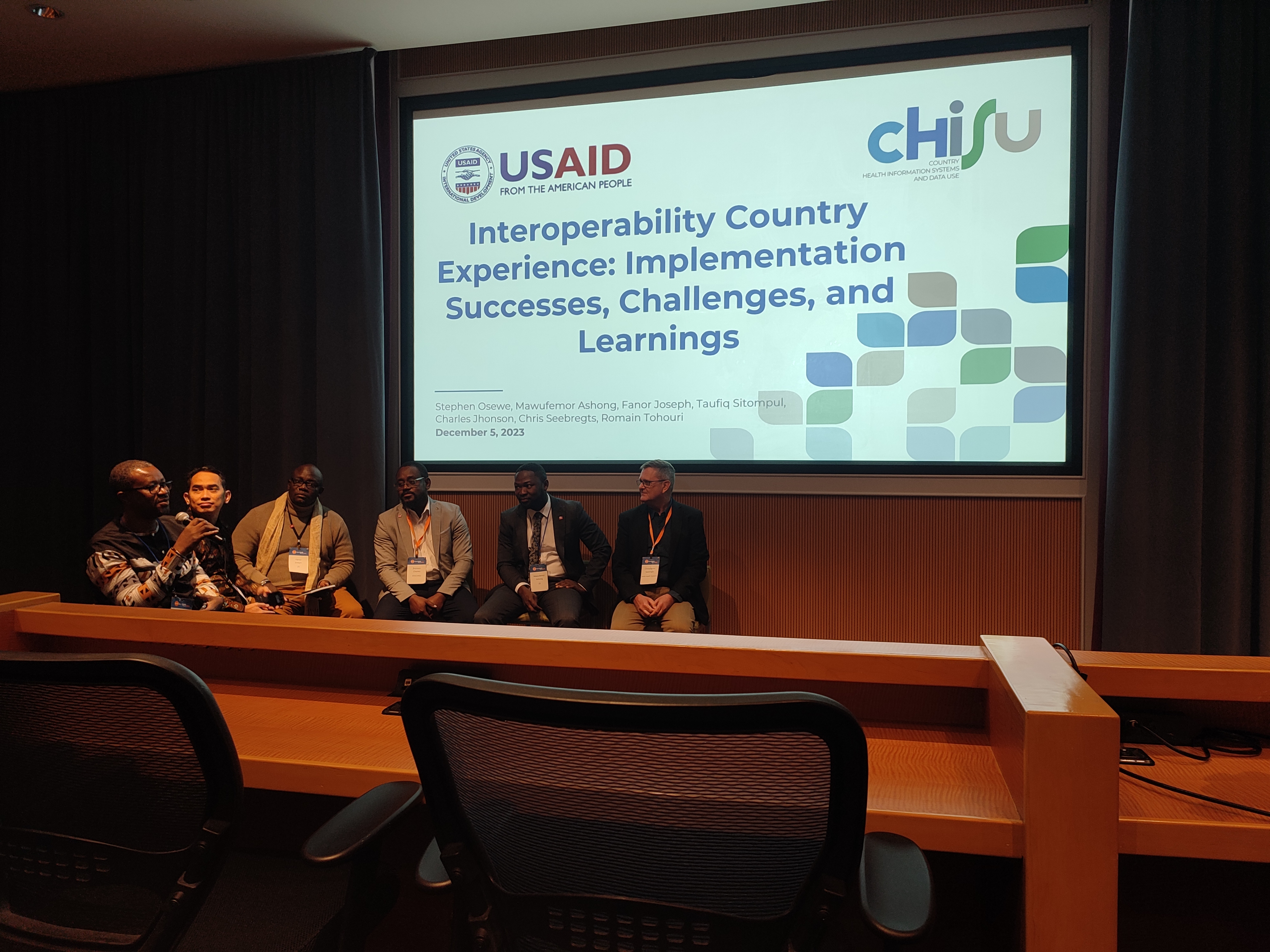 Six men presenting at GDHF 2023 sit in front of a presentation projection. The title slide reads: "Interoperability Country Experience: Implementation Successes, Challenges, and Learnings