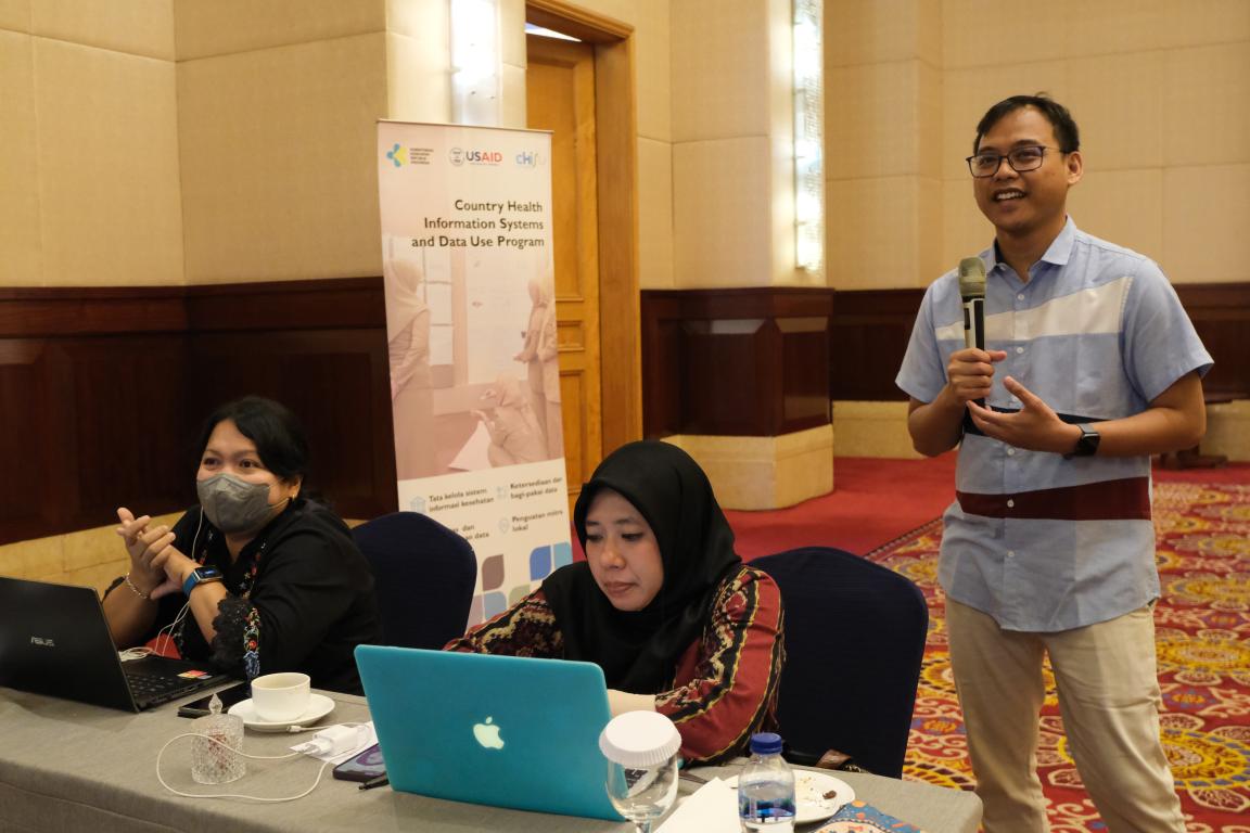 A man stands and speaks at a data quality assurance workshop and two women look at their laptops.