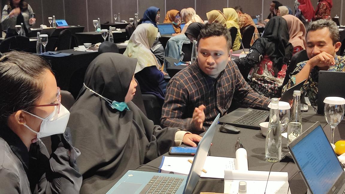 A man speaks to two women sitting at a table with laptops in front of them.