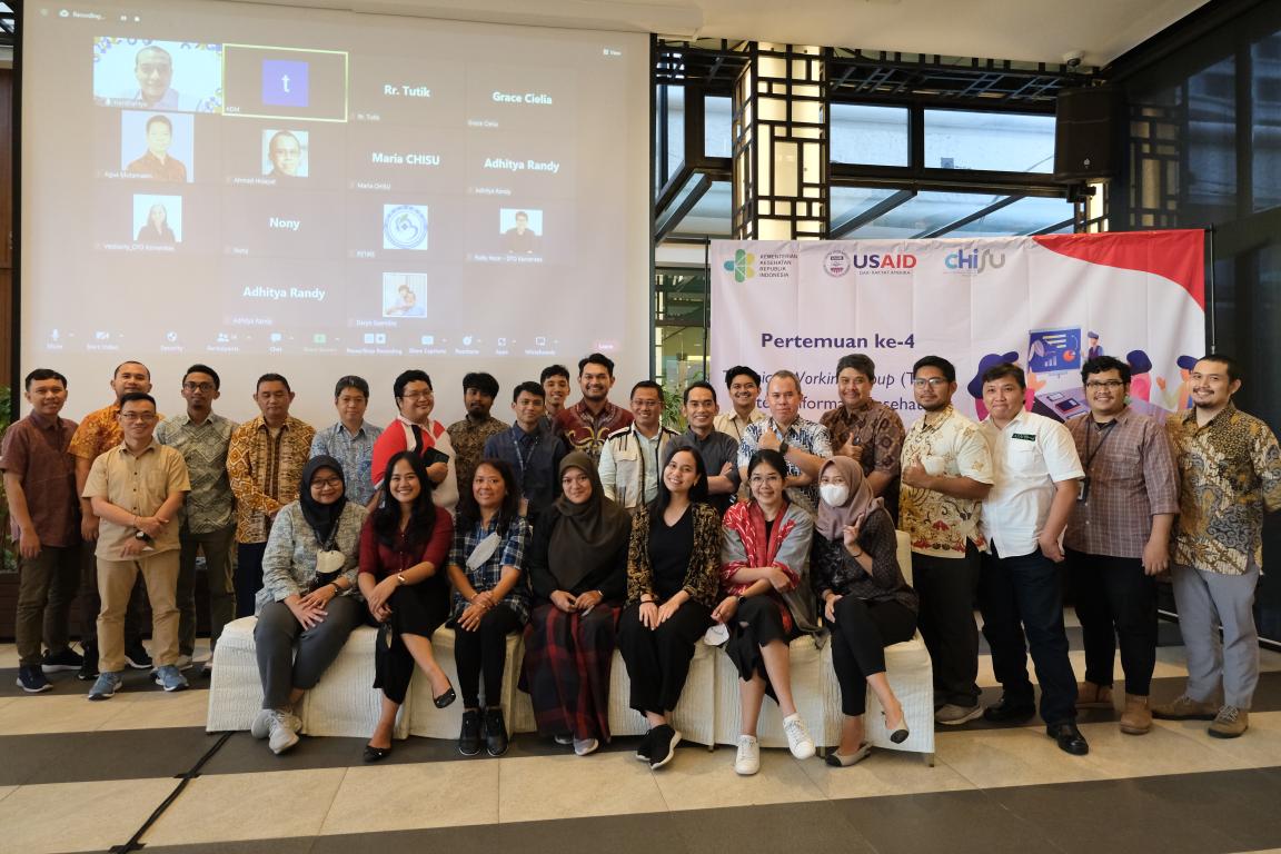 A group of people stands in front of a banner and projector screen with a Zoom window open.