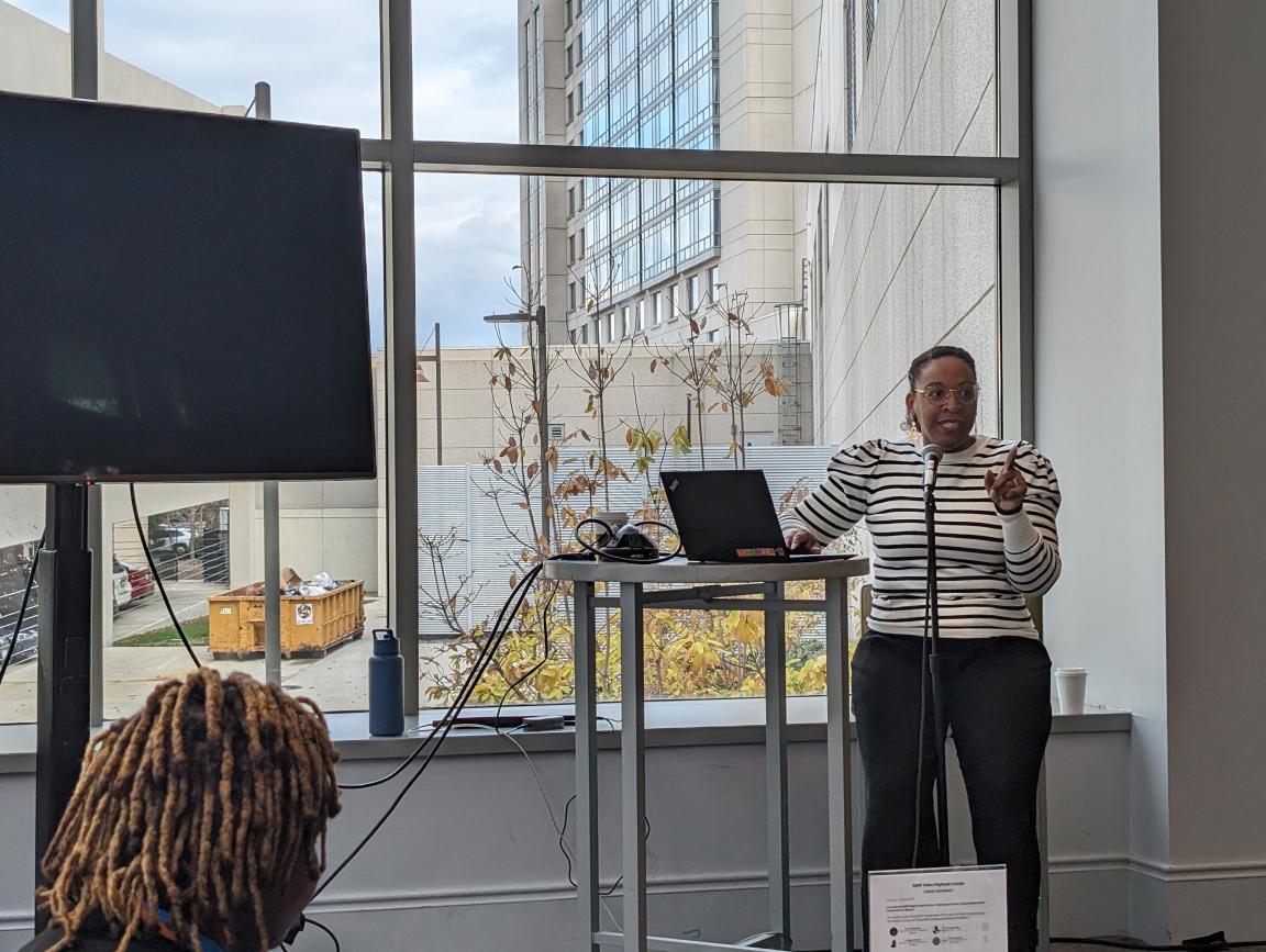 A woman stands at a podium with a laptop and speaks to a group of people.